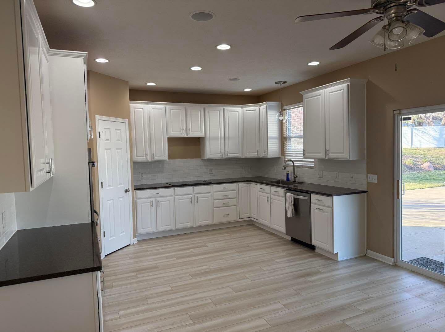 Bright empty kitchen with white cabinets, gray countertops, wood-look floors, and a ceiling fan.