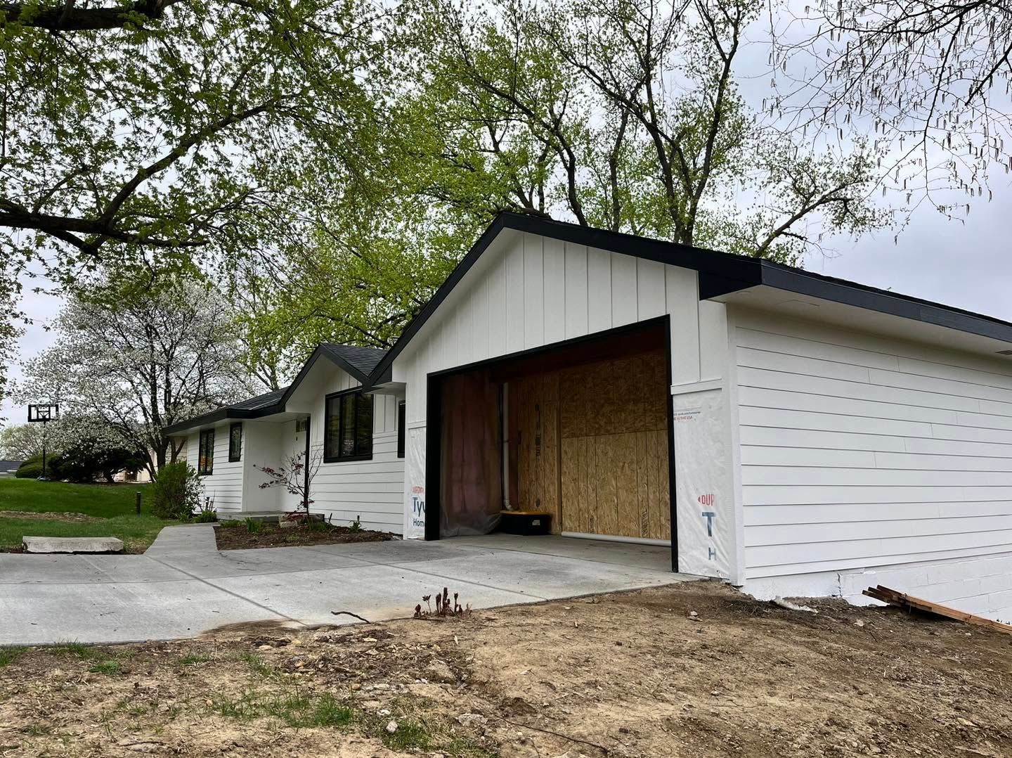White house with attached garage under construction, viewed from the driveway with trees overhead