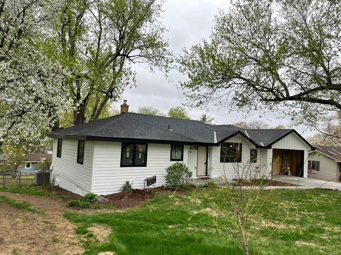 White house with black roof and windows, surrounded by trees and grass in a rural yard.