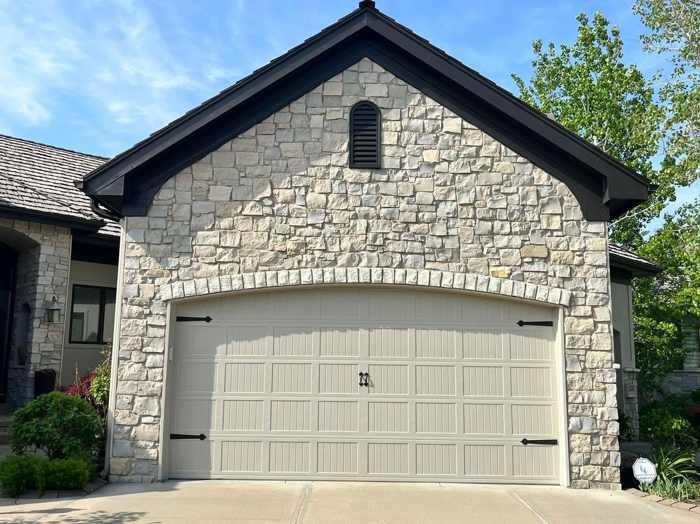 Stone garage with beige double door and dark shingled roof in a suburban setting