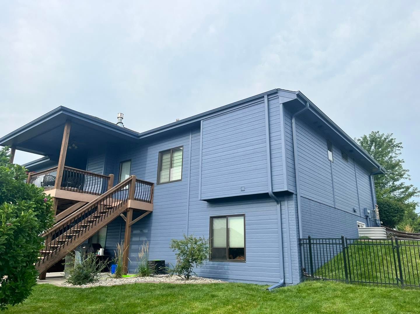 Blue two-story house with a wooden staircase and porch, set on a grassy yard under a cloudy sky