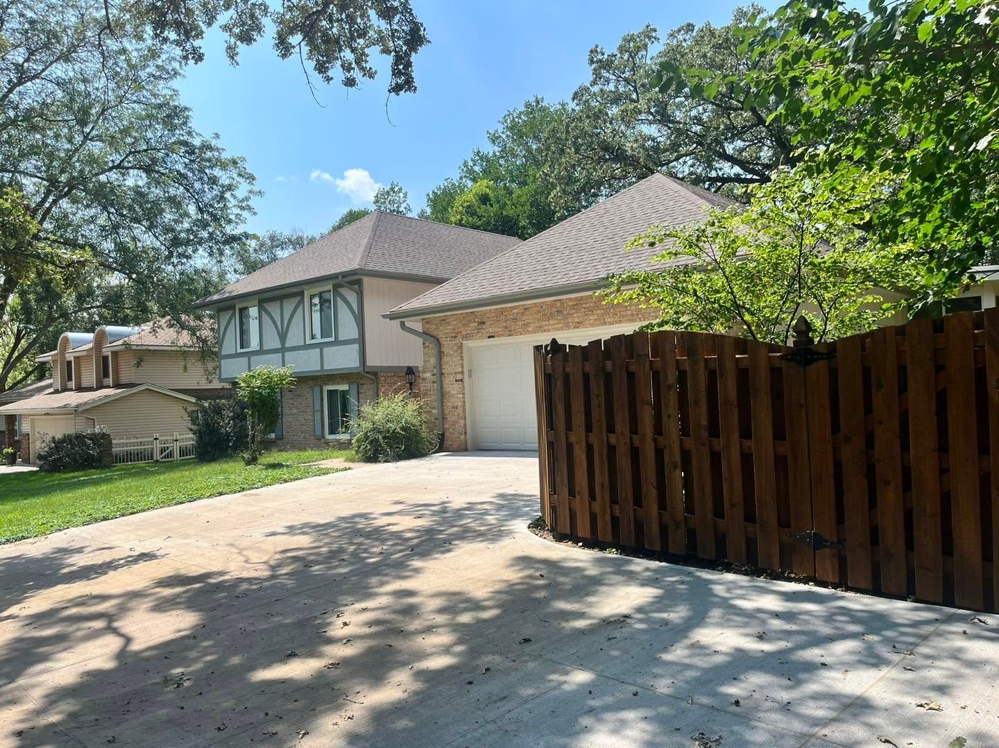 Suburban house with two-car garage, paved driveway, and wooden fence under trees on a sunny day