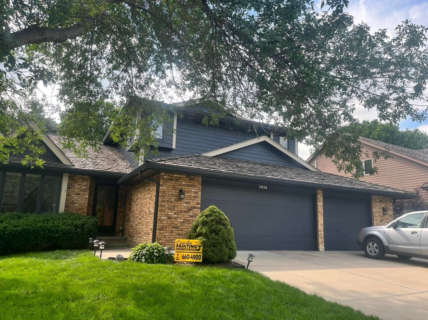 Suburban house with dark roof and garage, green lawn, tree shade, and parked silver car