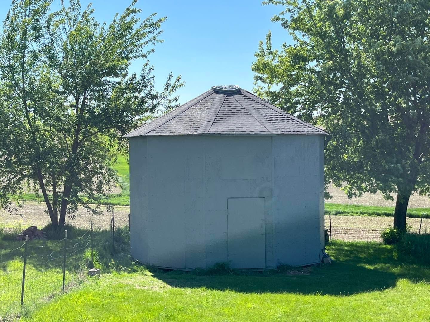 Small gray shed with a dark shingled roof, centered in a grassy yard with trees behind it.