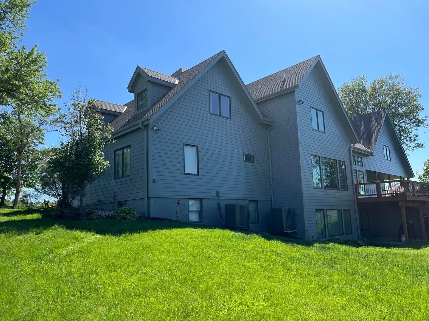 Gray house with steep roofs and a deck, surrounded by a sunny green lawn and trees.