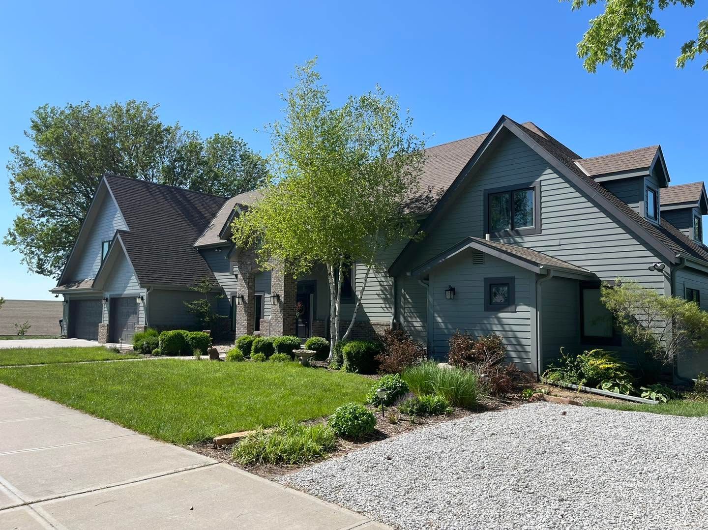 Suburban house with gray siding, green lawn, and gravel driveway on a sunny day