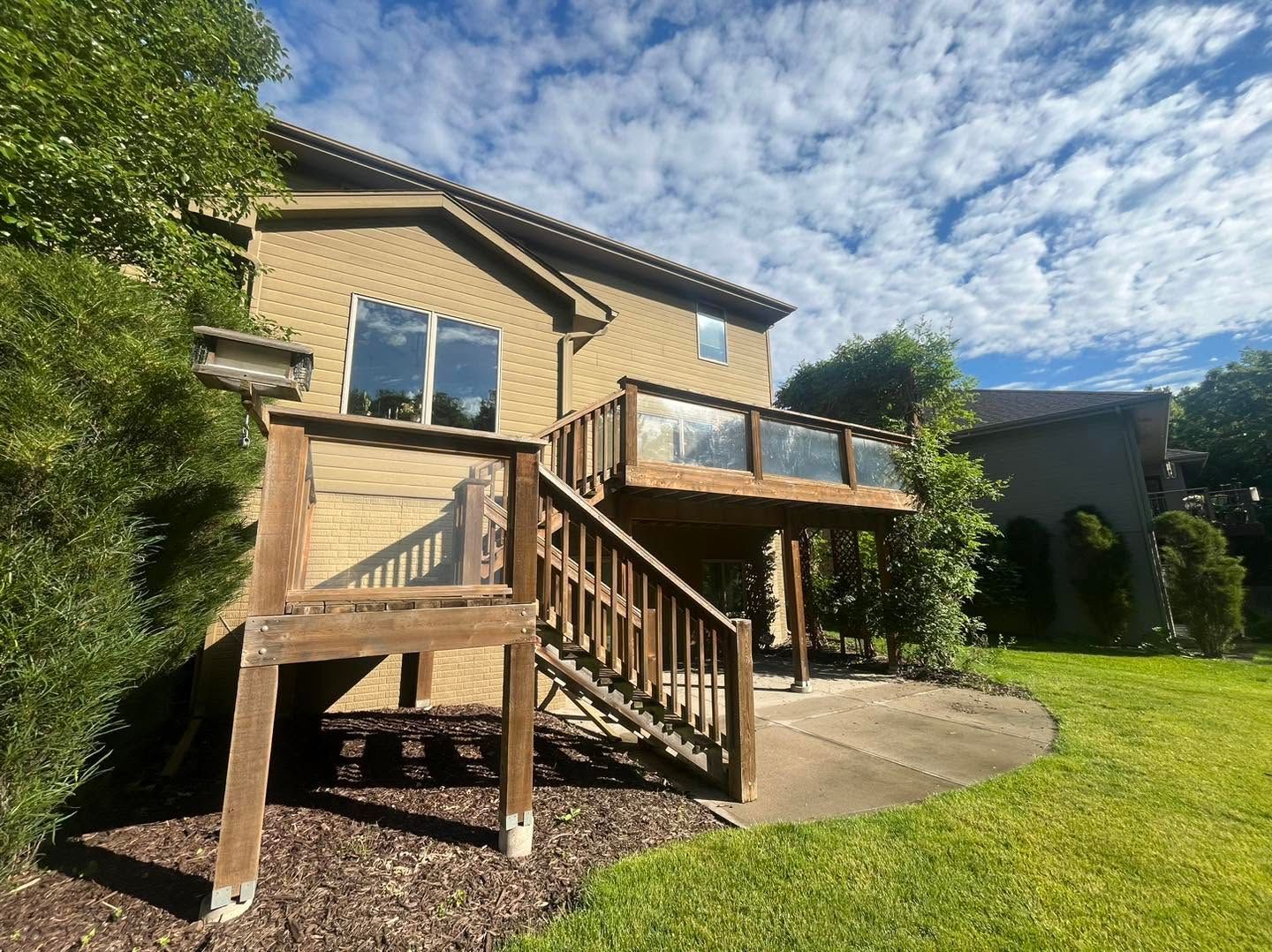 Backyard view of a tan house with a wooden deck, stairs, and green lawn under a blue sky