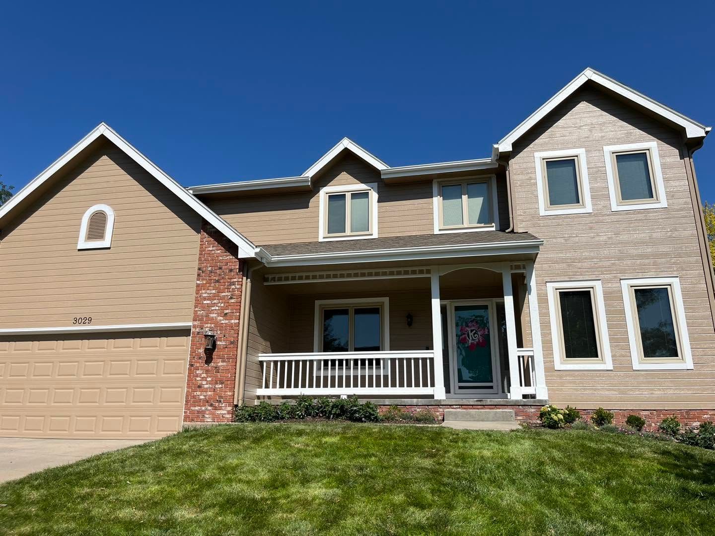Suburban two-story house with beige siding, brick accents, attached garage, and a front porch