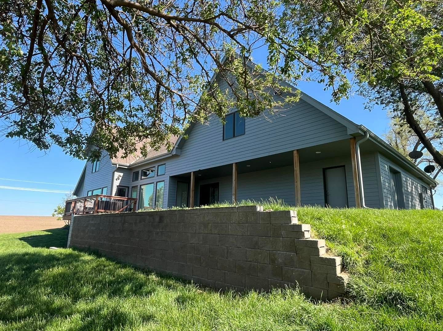 Blue farmhouse with porch and stone retaining wall on a grassy hillside under a sunny sky