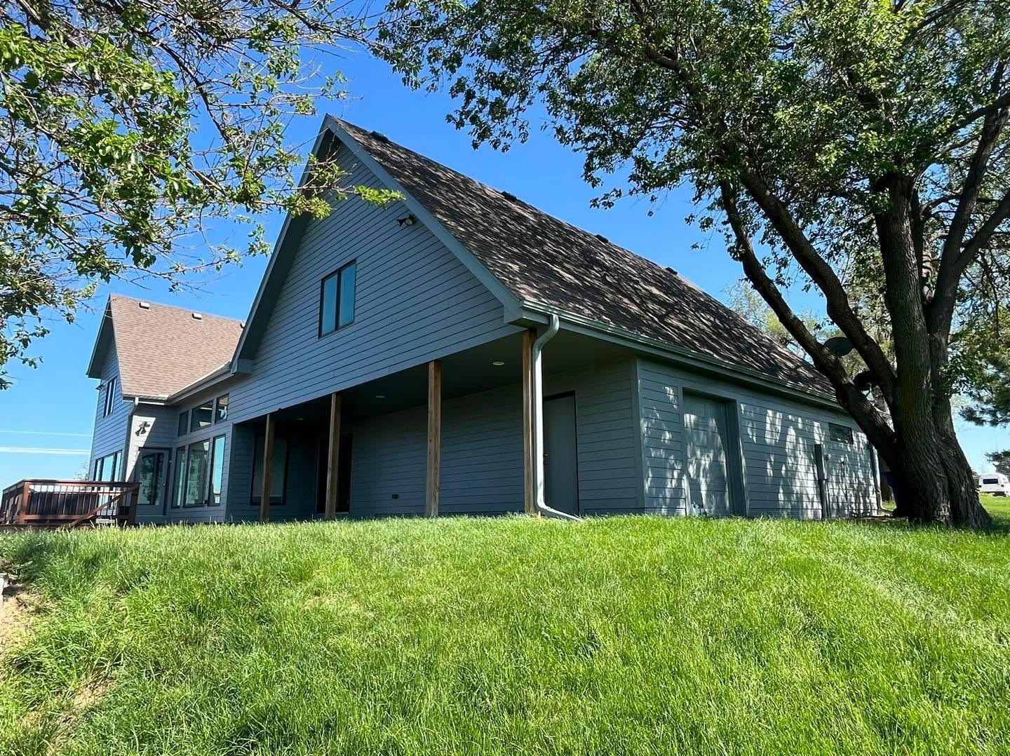 Large blue house with a porch on a sunny grassy hillside, framed by trees