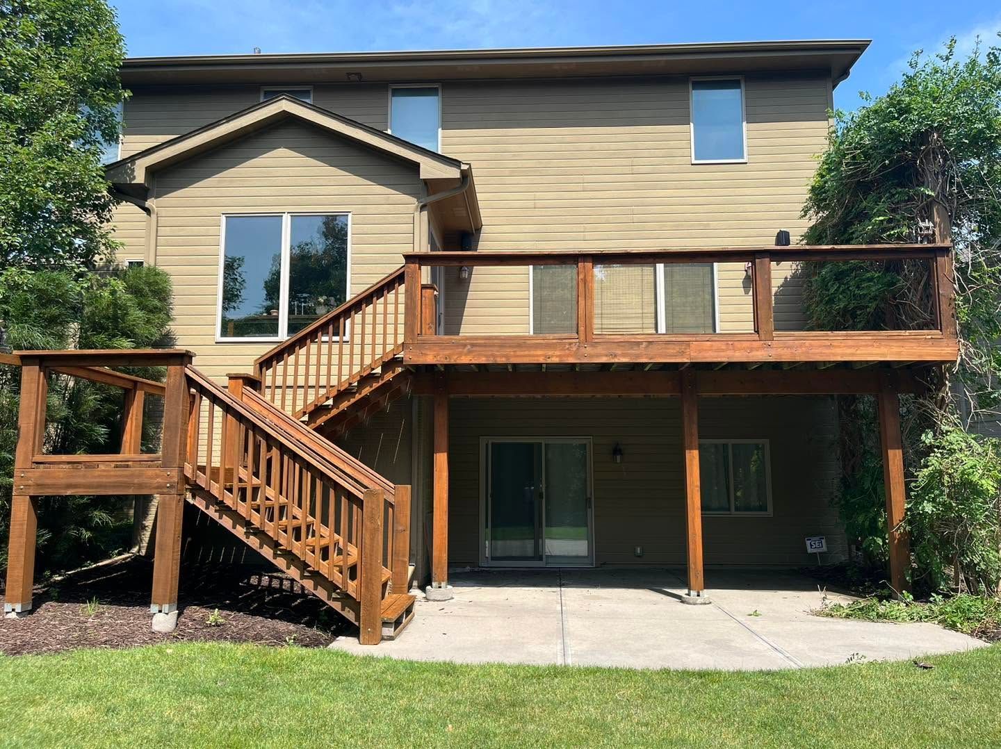 Back of a tan house with a wooden deck, stairs, and sliding doors leading to a patio and lawn.