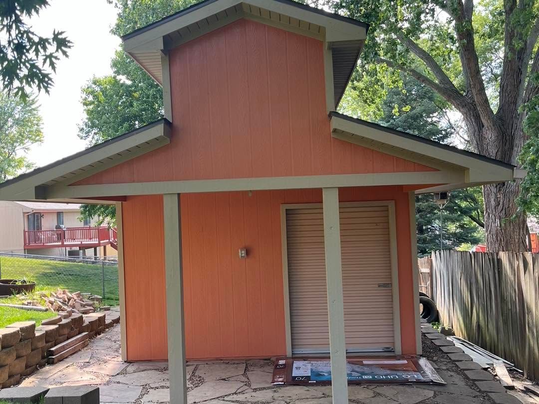 Small orange building with a porch, slanted roof, and closed door beside a fence and trees.