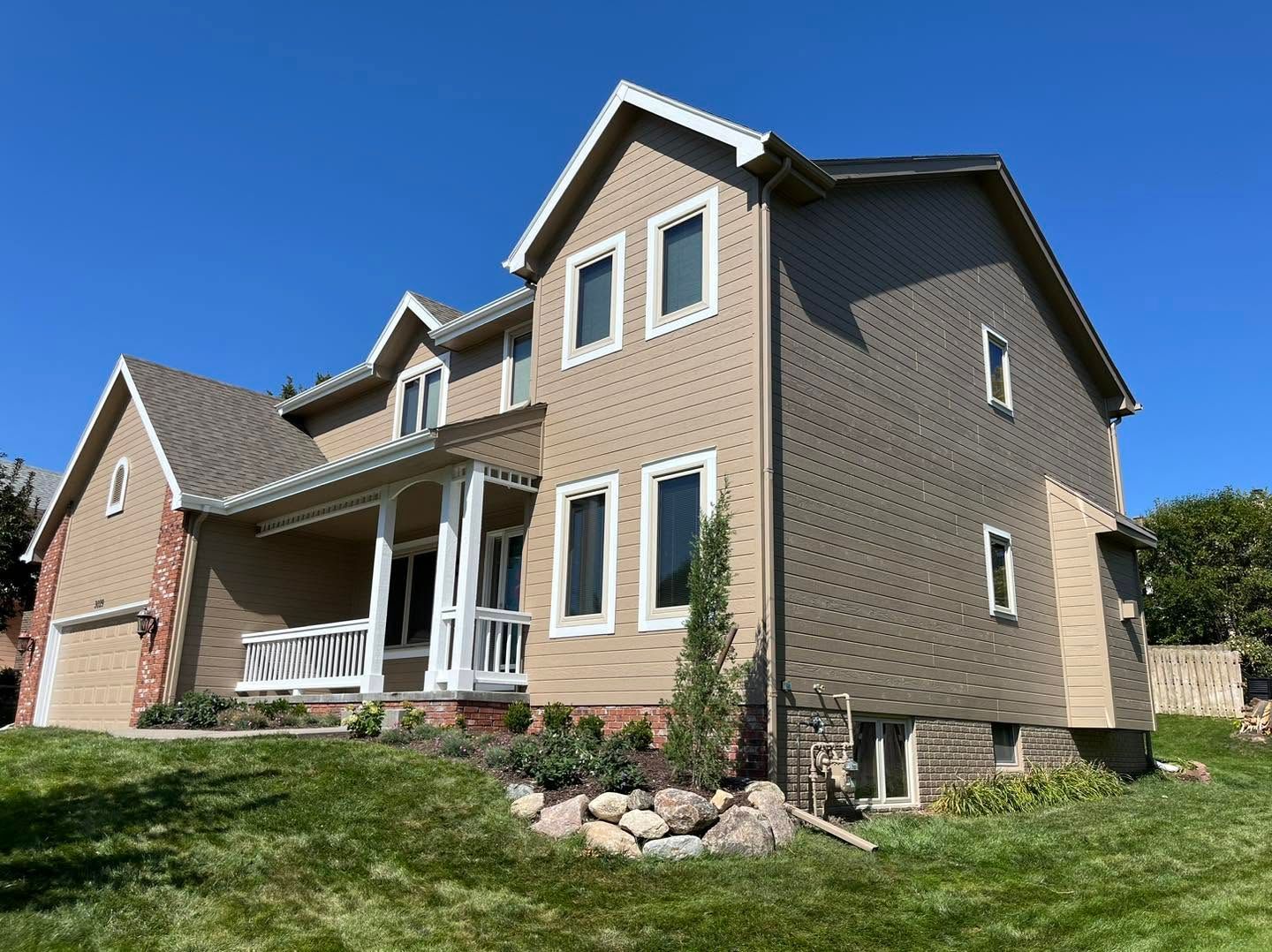 Suburban two-story house with tan siding, brick accents, and a small front porch on a sunny lawn