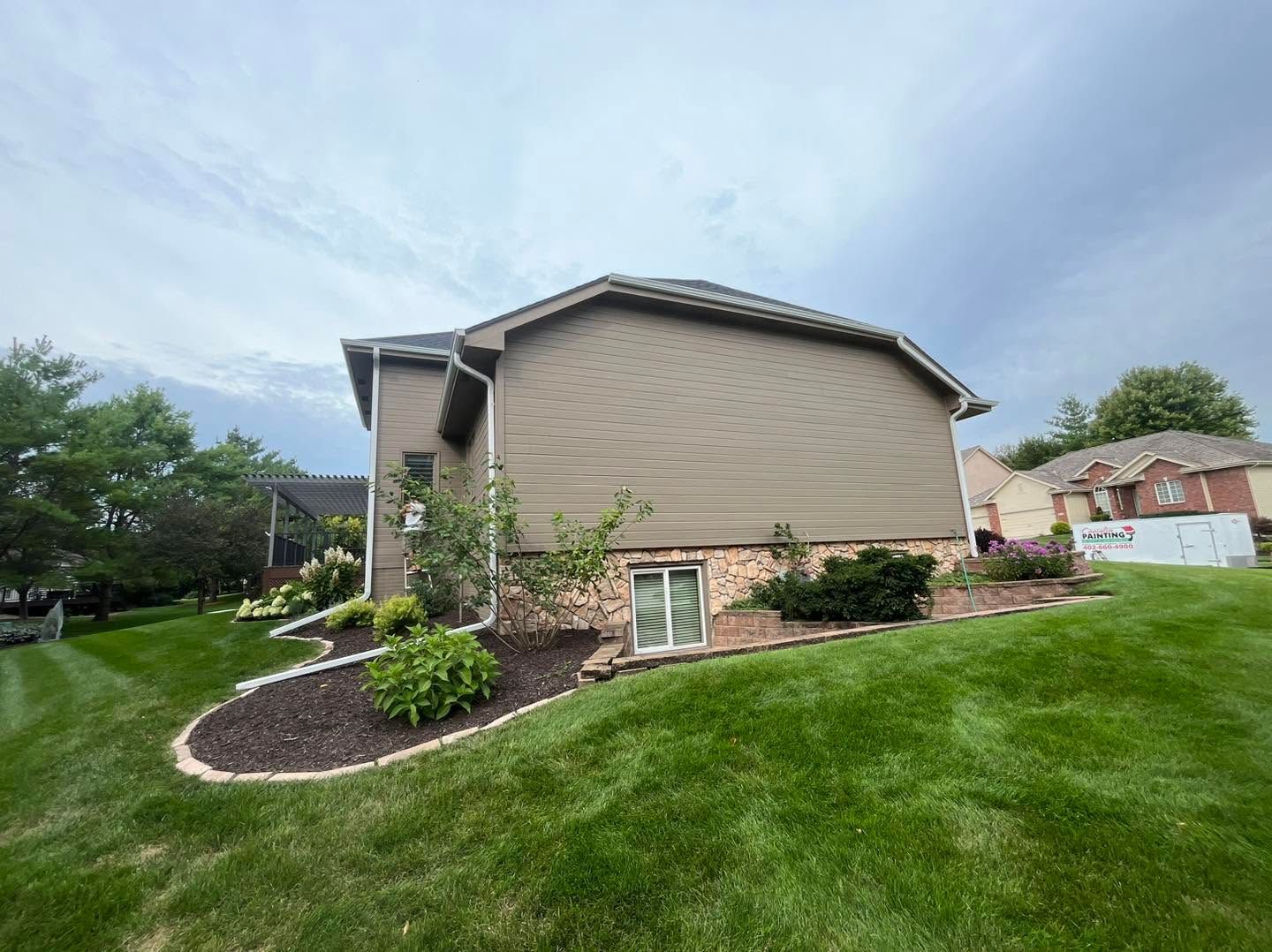 Suburban house exterior with beige siding, landscaped front yard, and green lawn under a cloudy sky