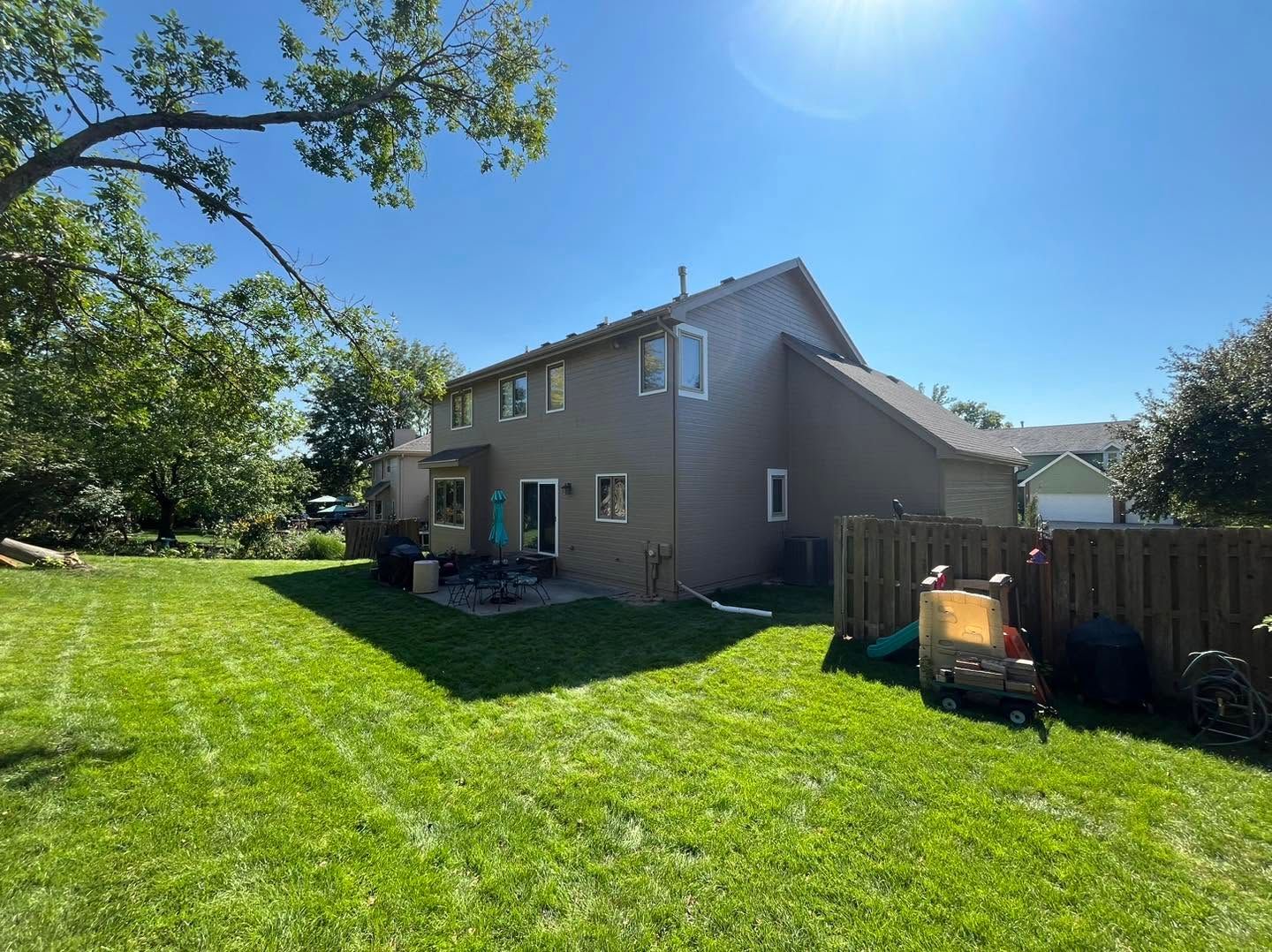 Sunny backyard with green lawn, two-story beige house, and wooden fence under a clear blue sky