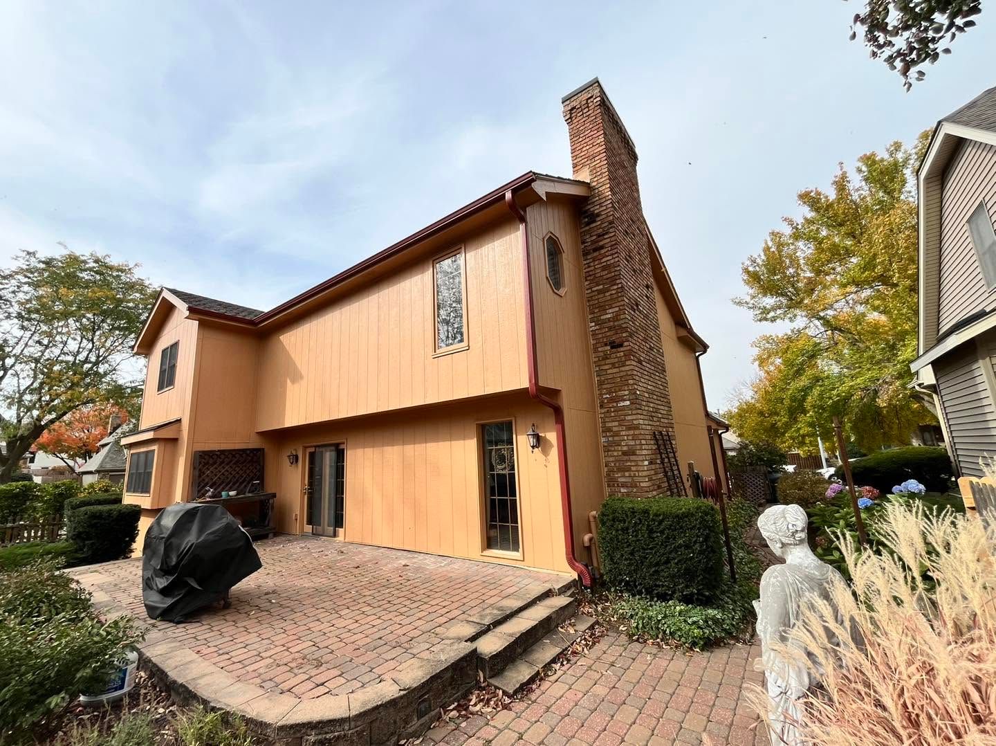 Two-story tan house with brick chimney, double garage, and paved driveway on a suburban street