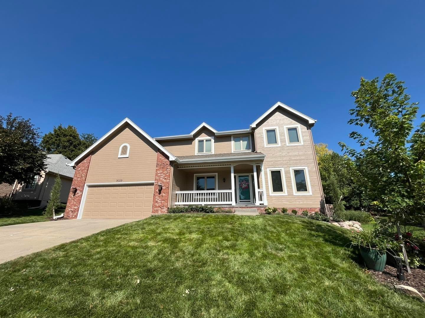 Suburban two-story house with tan siding, brick accents, and a large green lawn under a blue sky