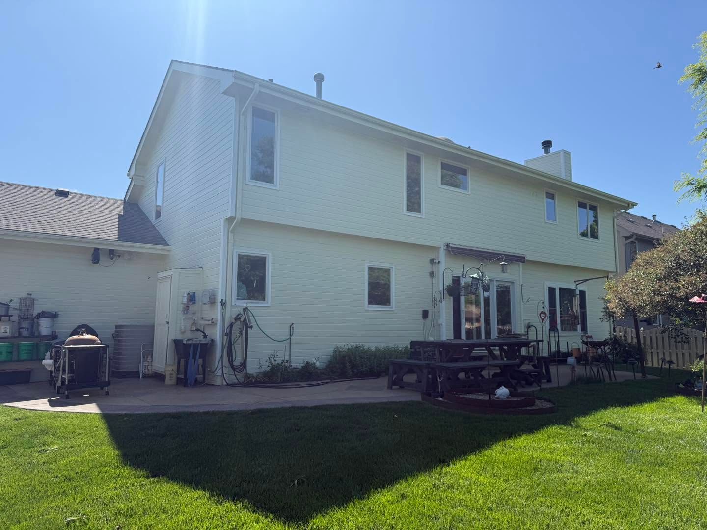 Backyard of a two-story white house with green lawn, patio furniture, and grill in sunlight