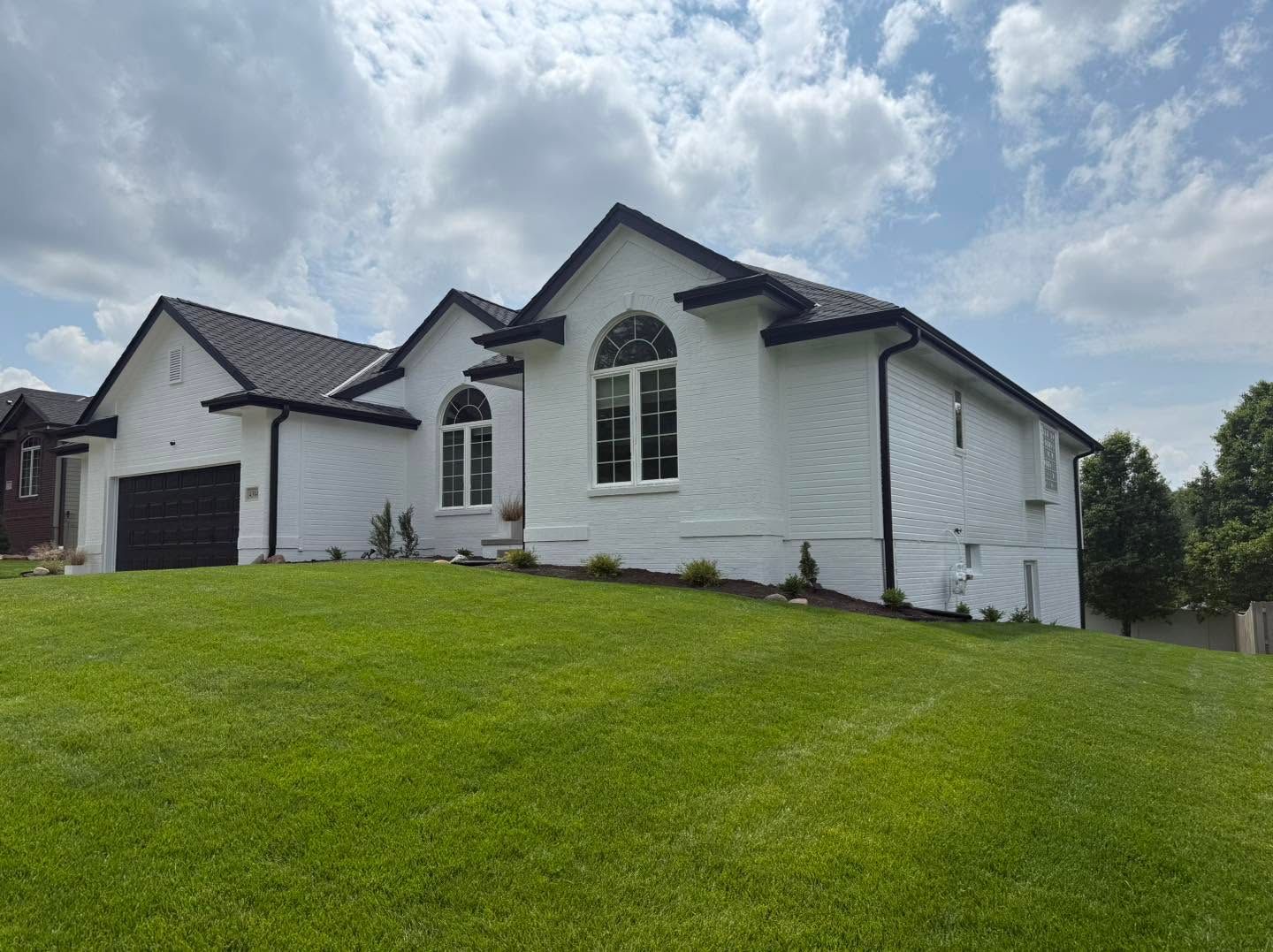 White single-story house with black roof and garage on a green sloped lawn under cloudy sky