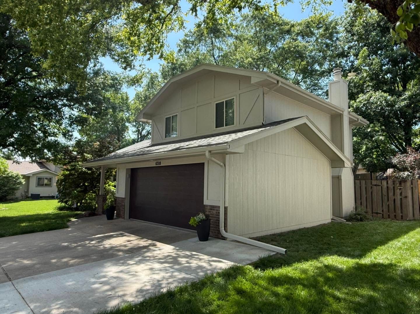 Two-story beige house with a dark garage door, driveway, and green lawn under trees