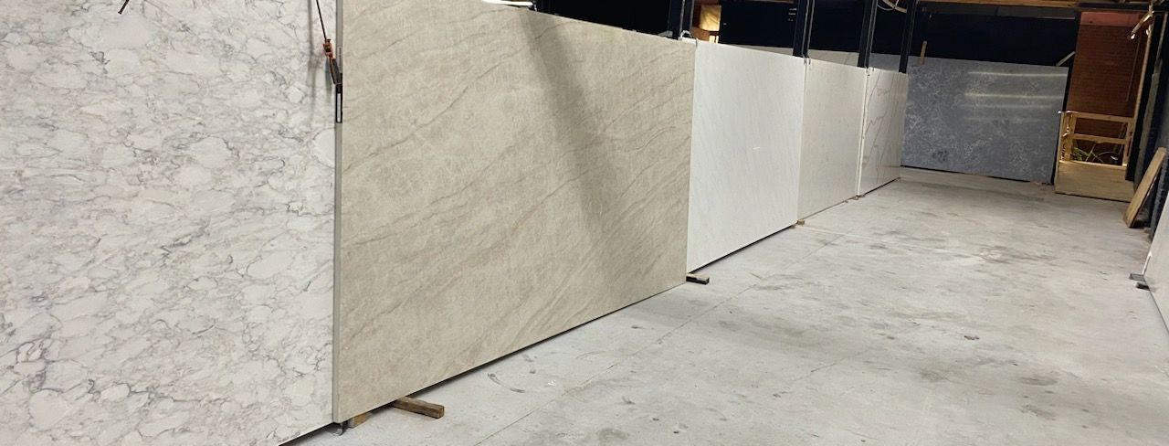 Large stone slabs of varying colors, possibly granite or marble, standing upright against a concrete floor. The slabs are indoors, likely in a showroom or storage area.