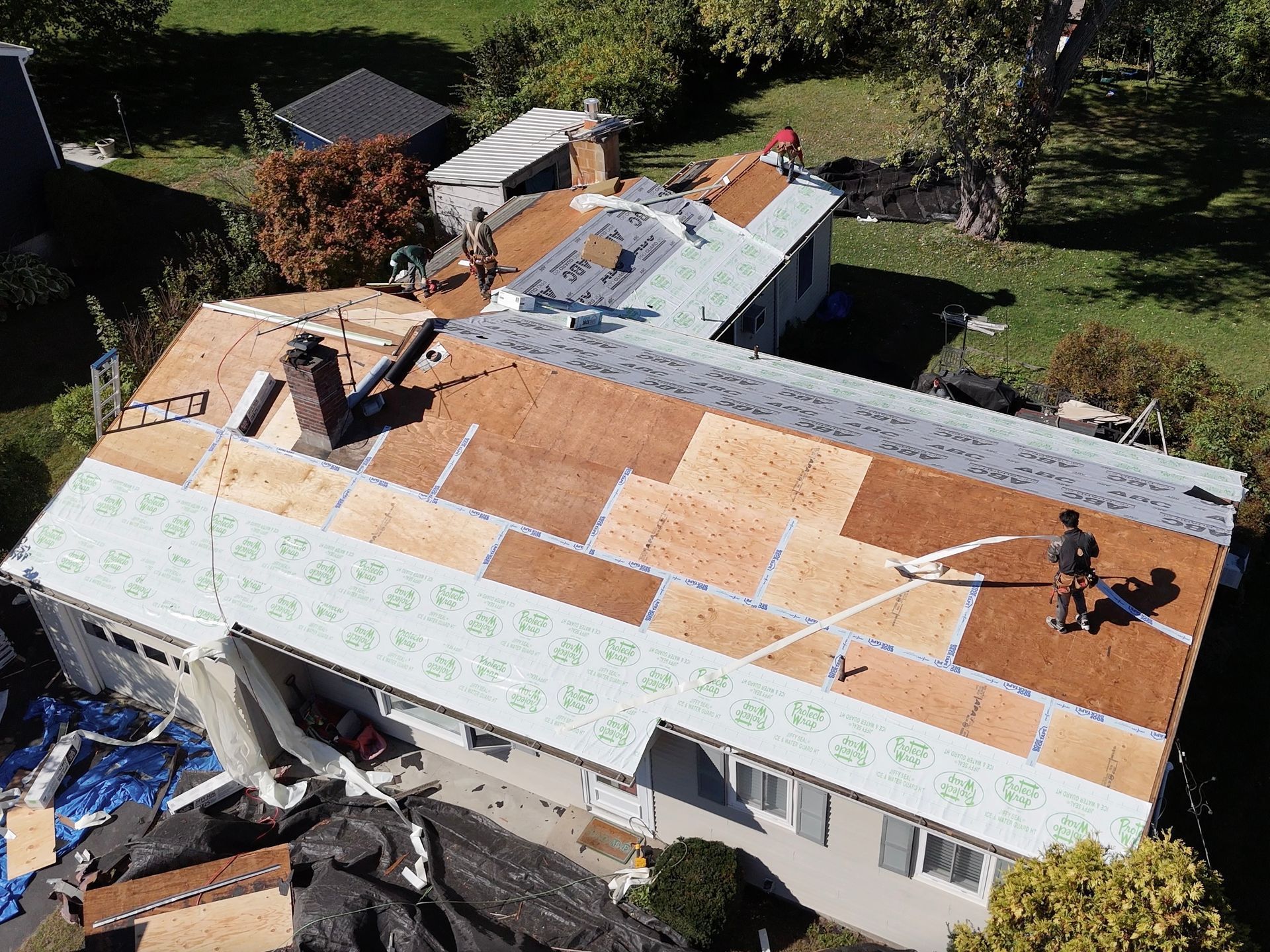 Roof being replaced on a house with workers, new wood, and blue tarps visible.