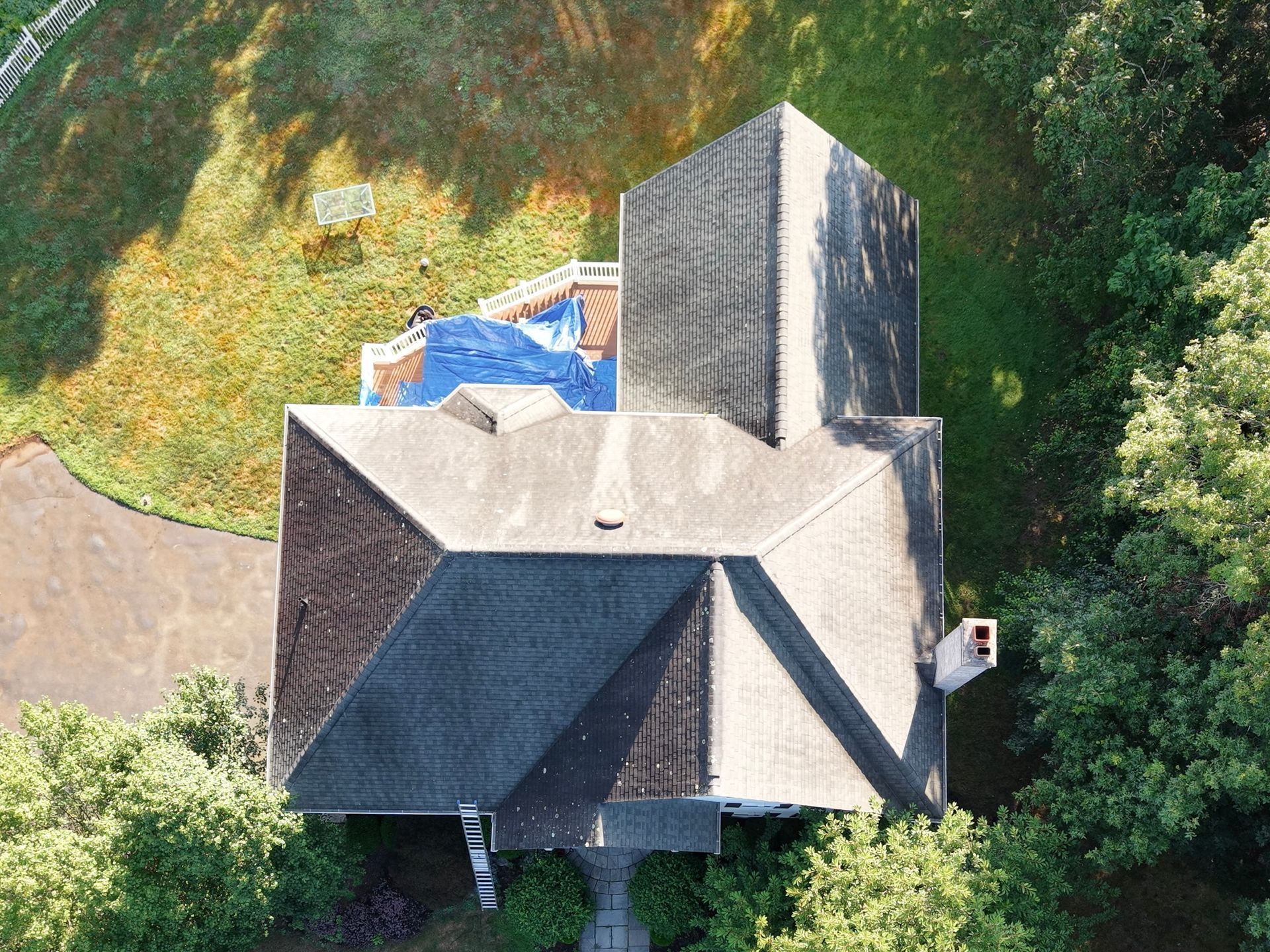 Overhead view of a house with dark gray roof, surrounded by green trees and lawn, with a blue tarp on the roof.