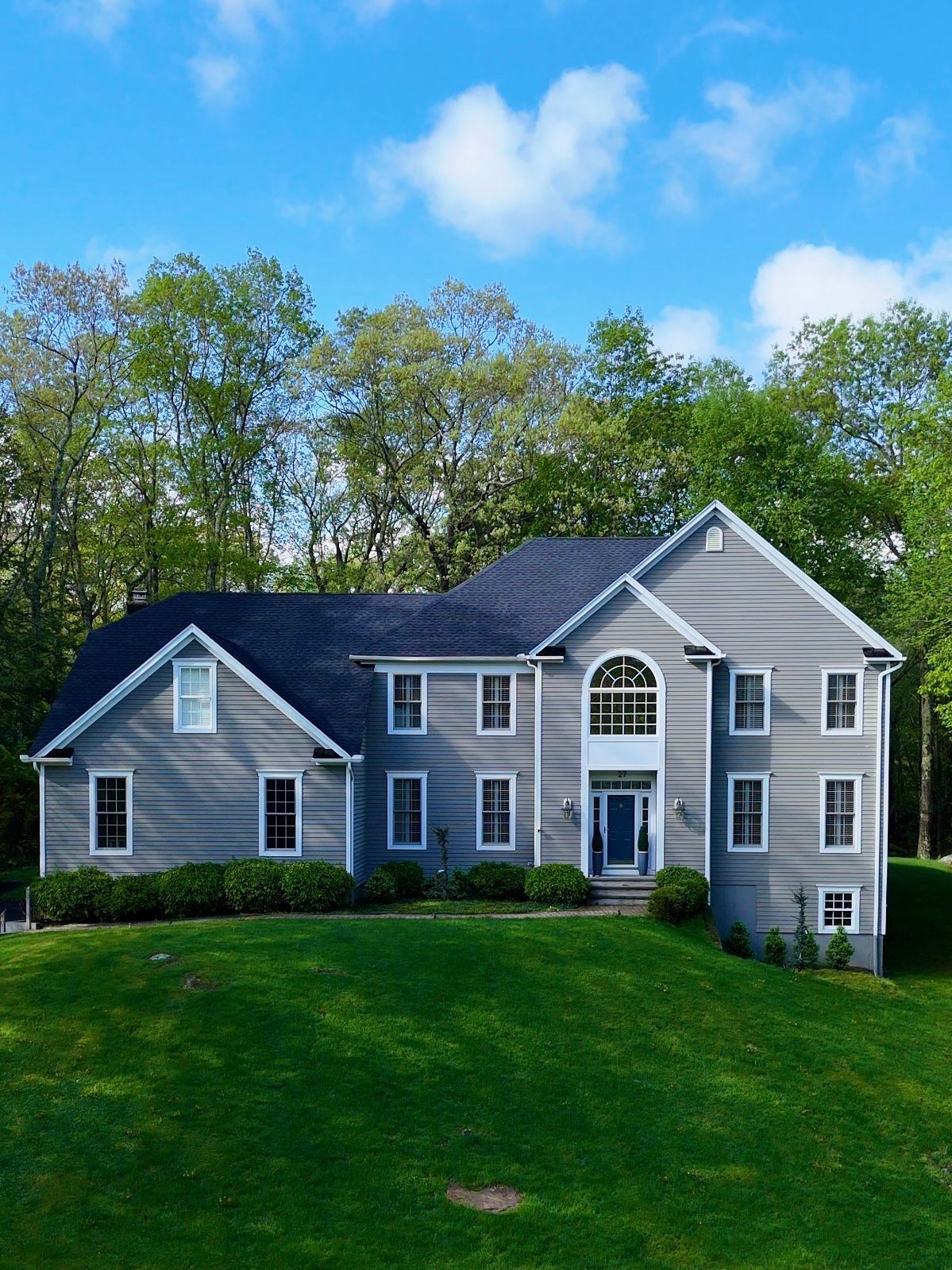 Gray two-story house with black roof, set on a green hill with trees and blue sky.