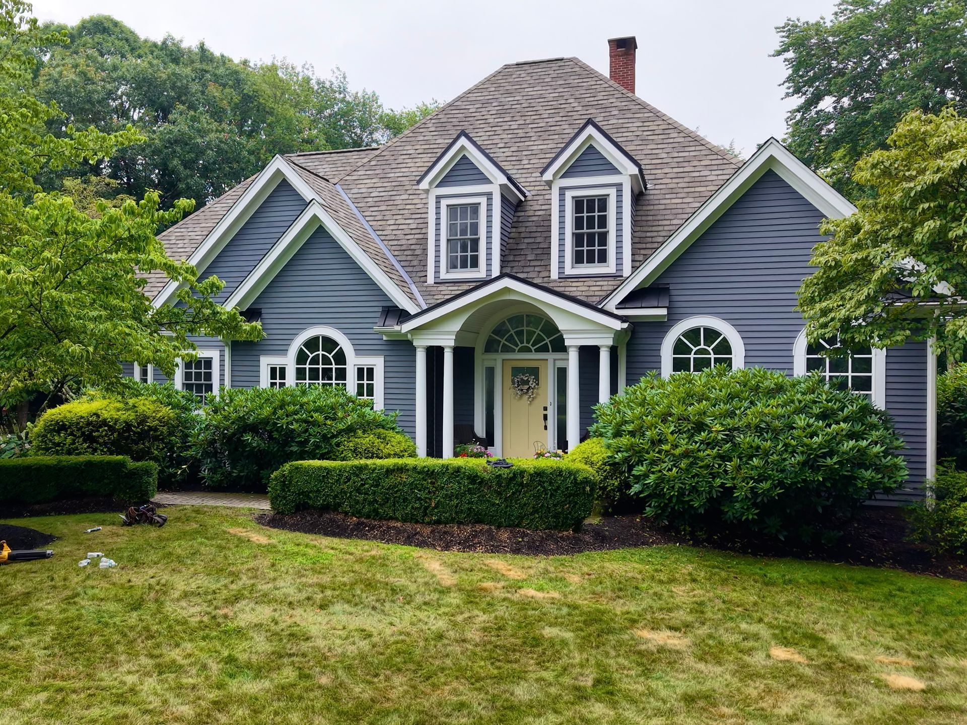Blue house with dormers, white trim, yellow door, surrounded by greenery and lawn.