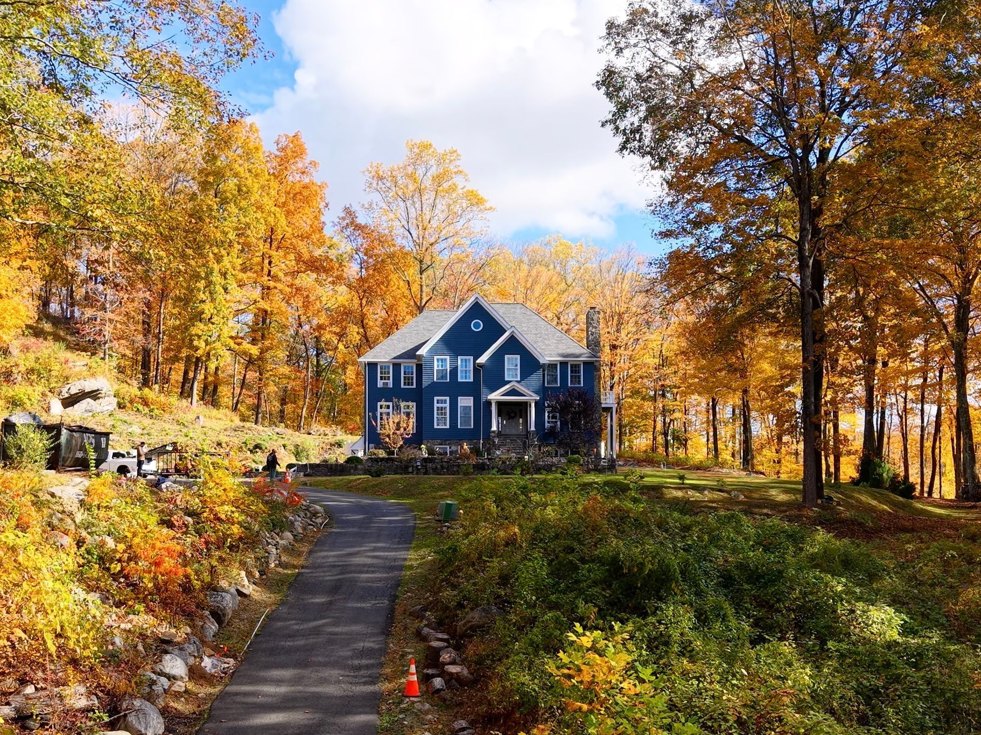 Blue house in a forest of trees with yellow and orange autumn foliage. A driveway leads to the house.