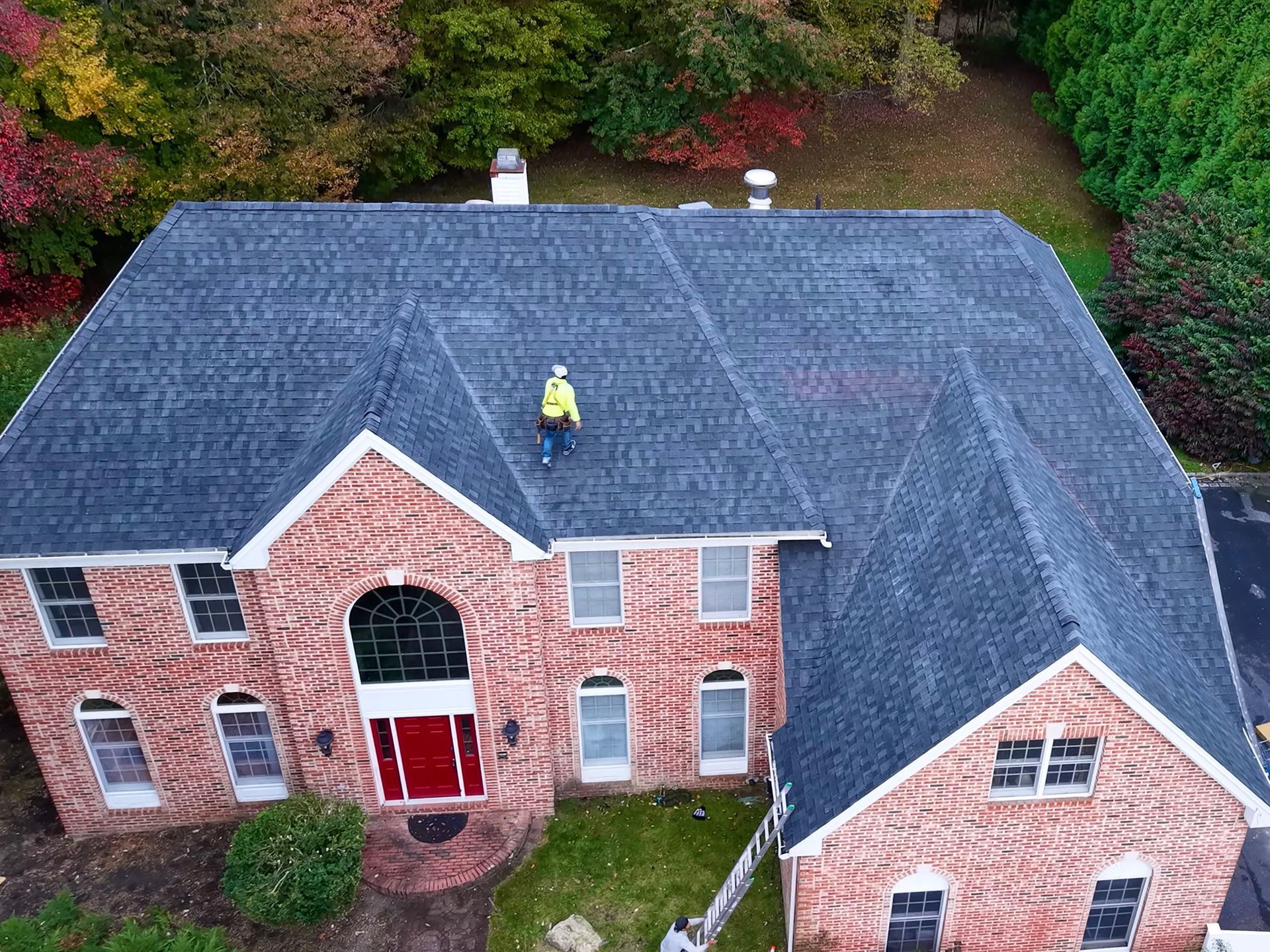 Man in yellow hard hat on roof inspecting. Red brick house with dark shingles, surrounded by trees.