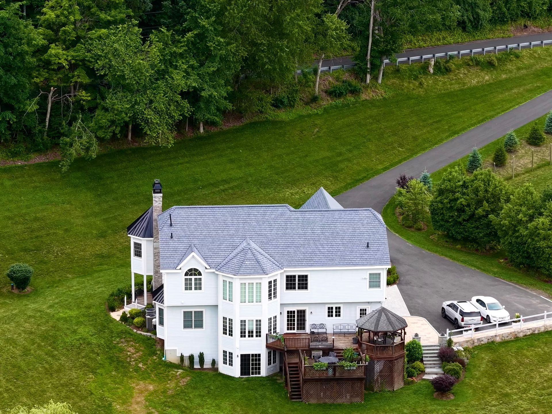 White house with gray roof, wooden deck, gazebo, and long driveway on a grassy hill. Two cars parked near the house.