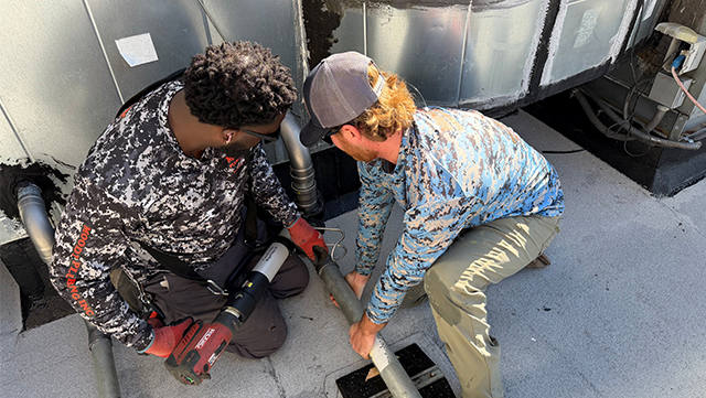 Man on rooftop inspecting with camera equipment, wearing yellow shirt. Bright sunny day.