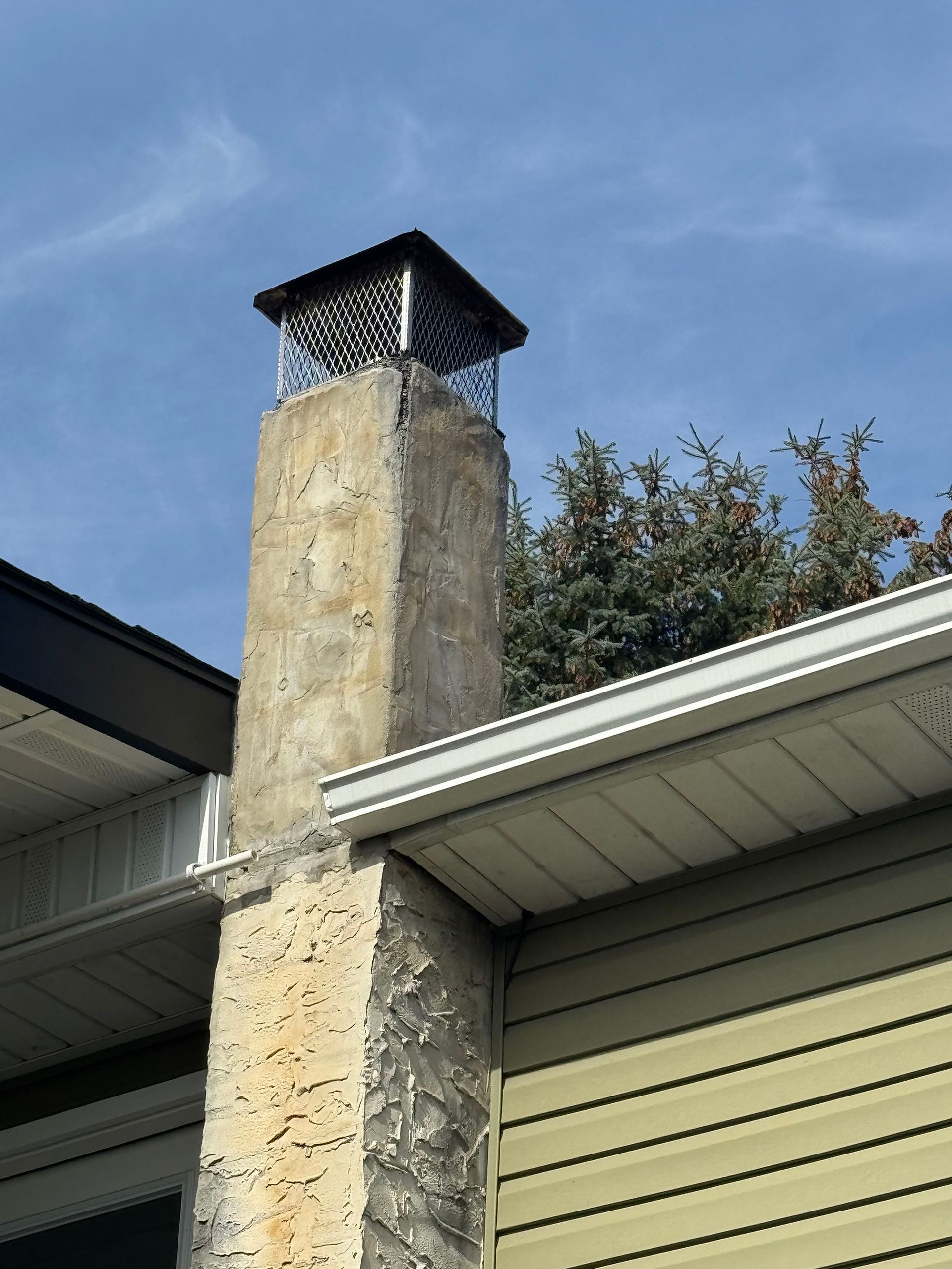 Chimney with a mesh cap, against a blue sky, attached to a house with yellow siding.