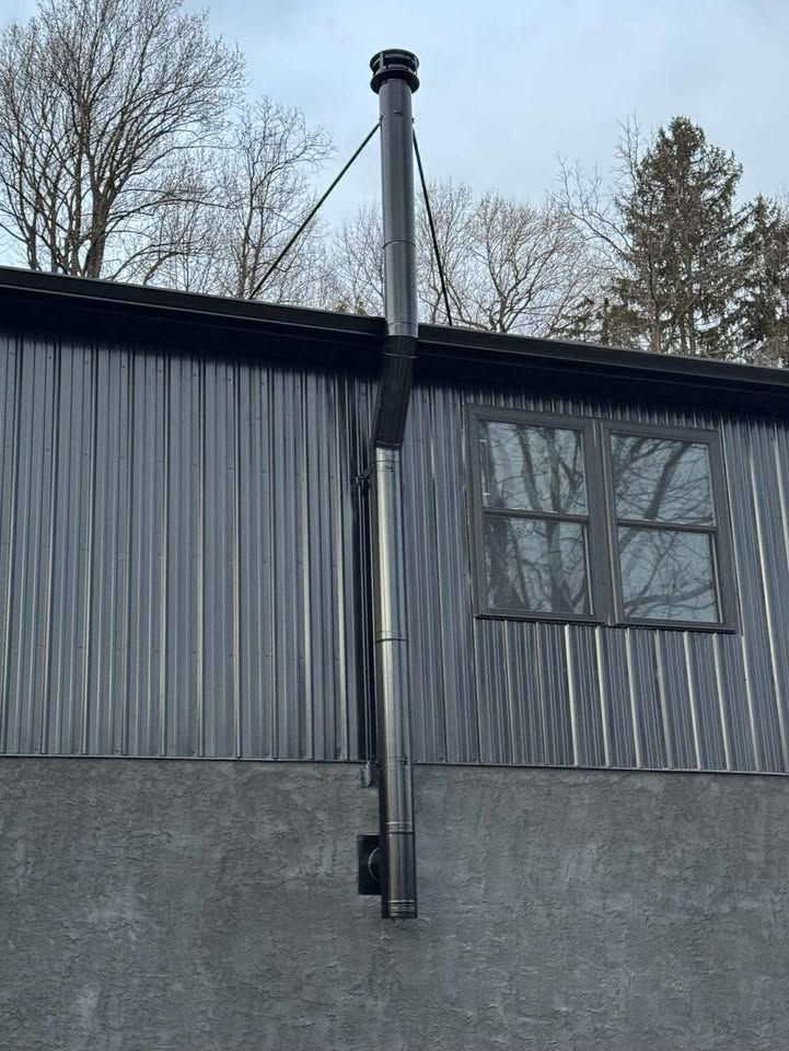 Black metal chimney on a corrugated gray building, secured by a cable. A window is visible.