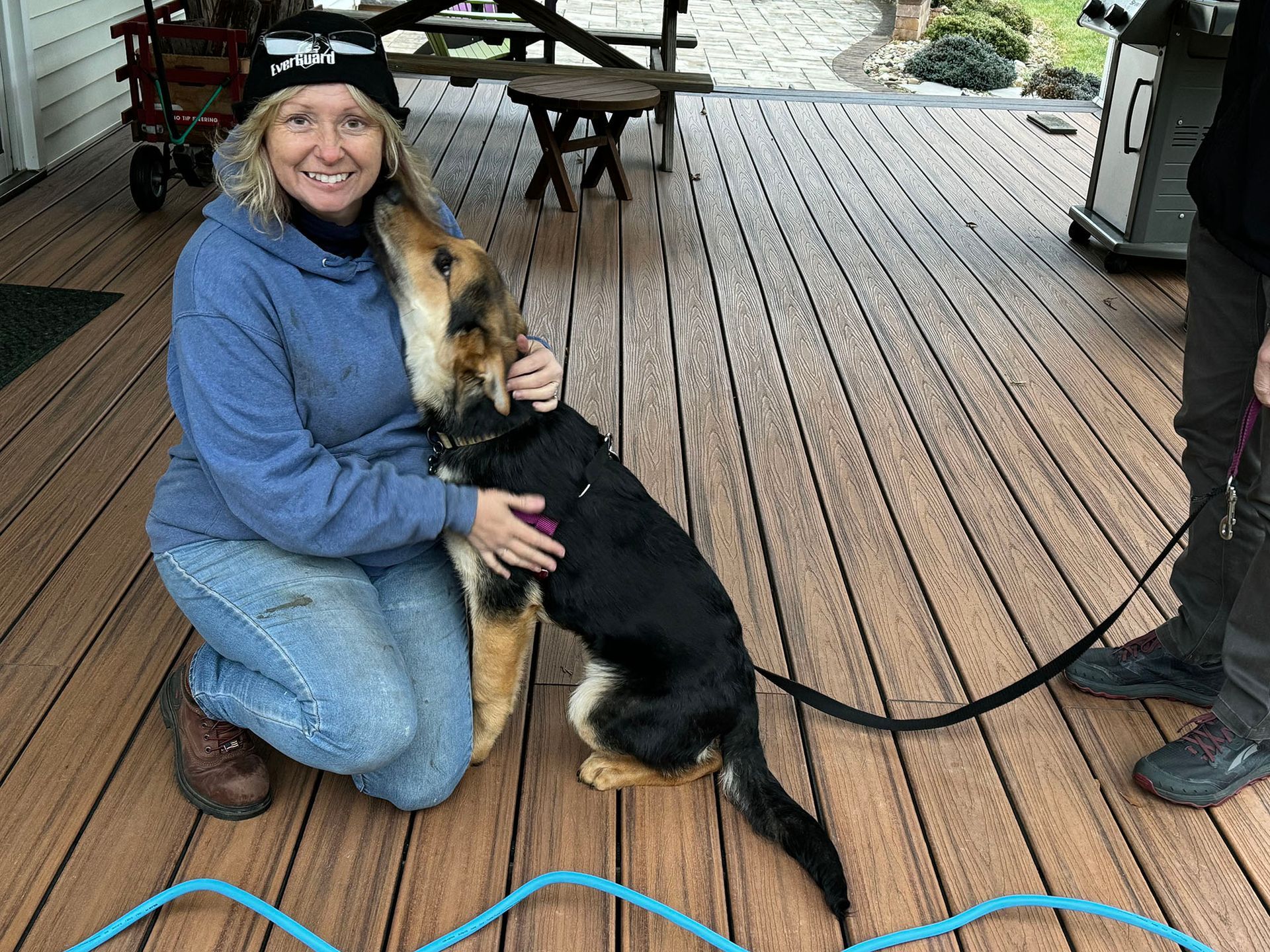 Woman kneeling, petting a black and tan dog on a wooden deck. Another person holds the dog's leash.