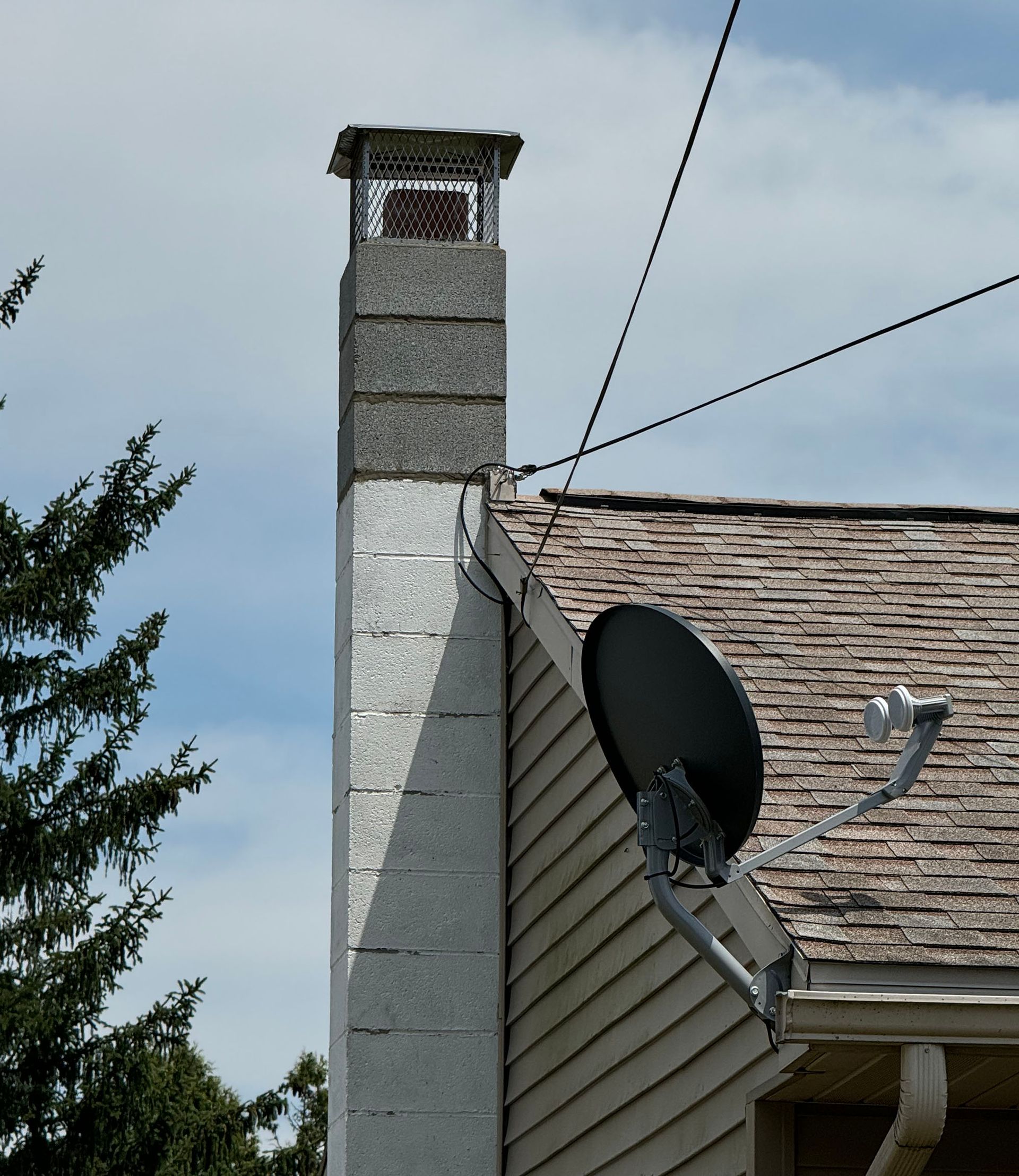 A tall, gray brick chimney with a metal cap rises from a roof. A satellite dish is mounted on the siding.