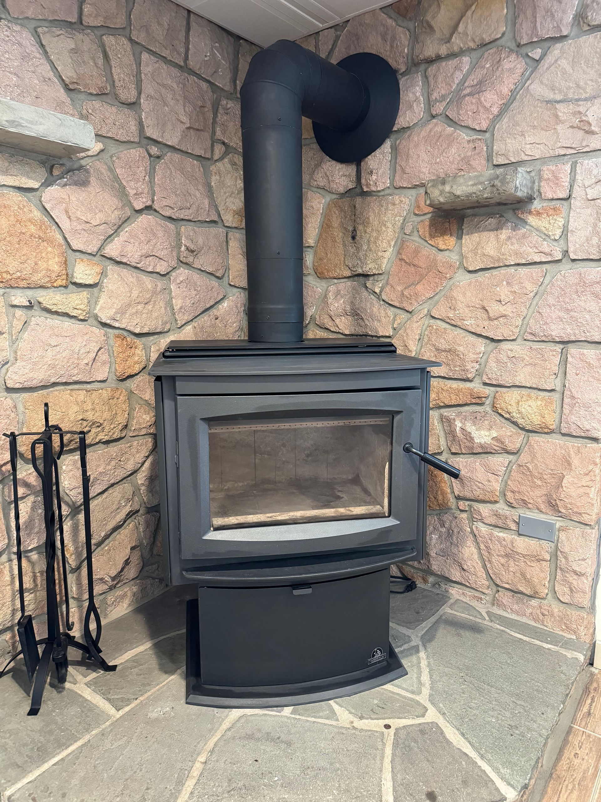 Black wood-burning stove with curved chimney pipe, set against a stone wall and stone floor.