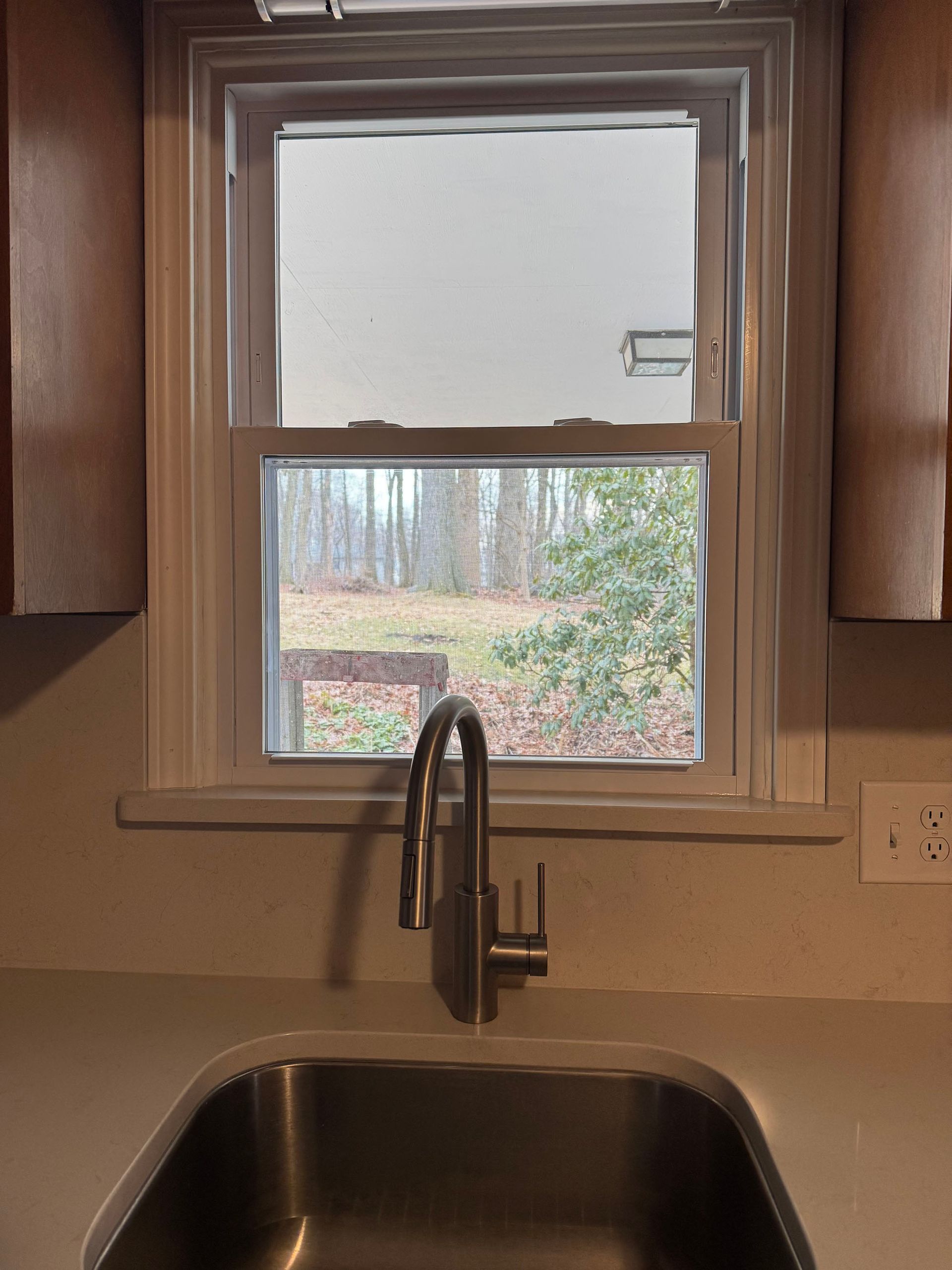 Kitchen sink with window above, showing a backyard view. A silver faucet is over the sink. Brown cabinets and a light countertop.