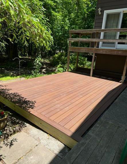 Wooden deck with railing attached to a brown house; surrounded by trees and stone patio.