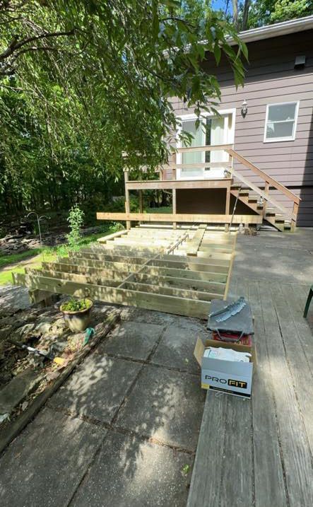 Deck construction in progress; wooden frame, partially built deck, gray patio, brown house, sunny outdoor setting.