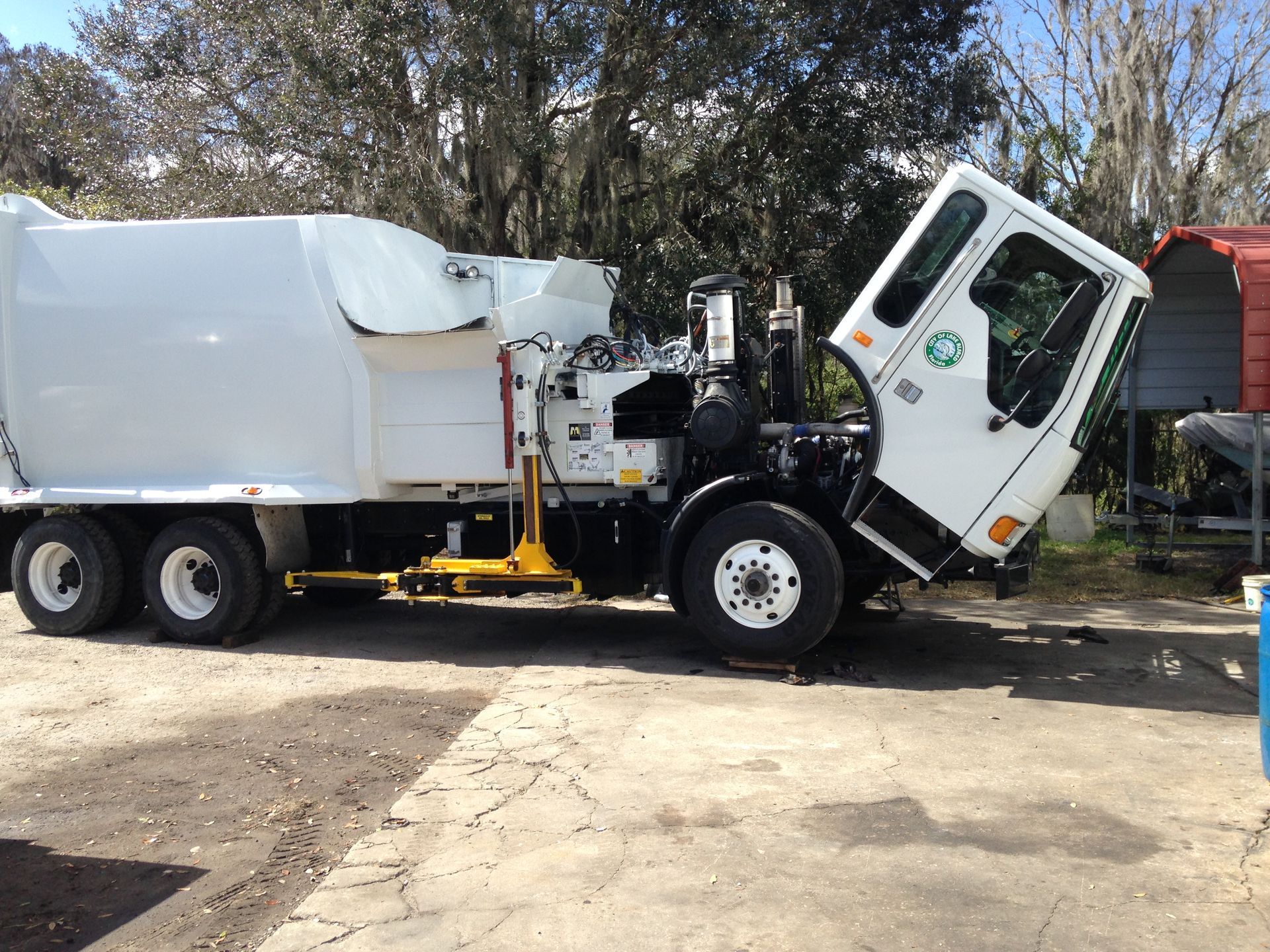 White garbage truck with tilted cab parked on a dirt road under trees