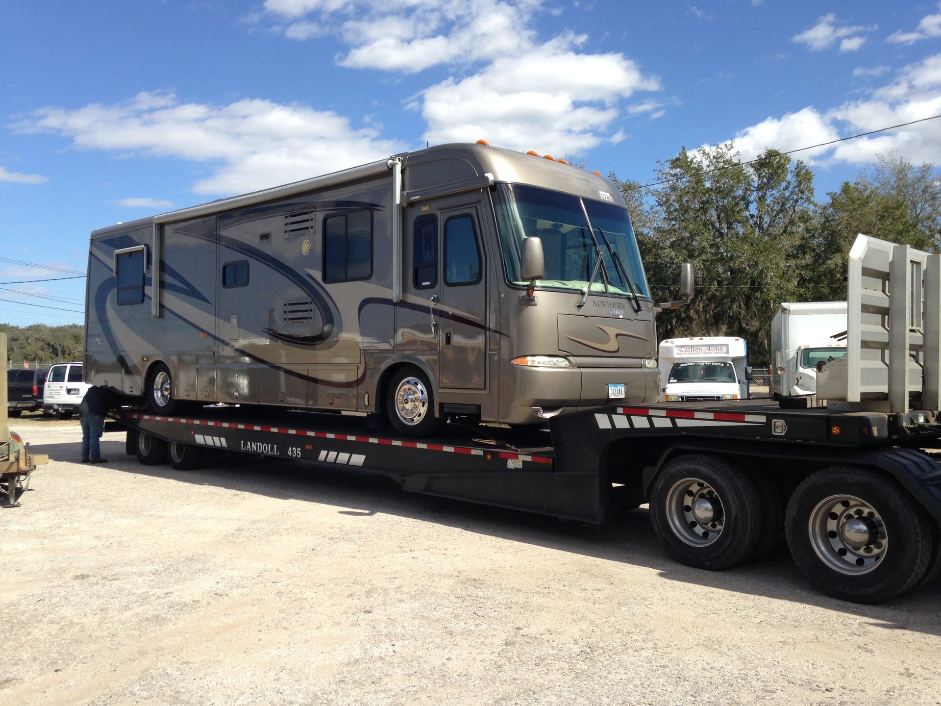 Beige motorhome secured on a flatbed trailer in a sunny parking lot