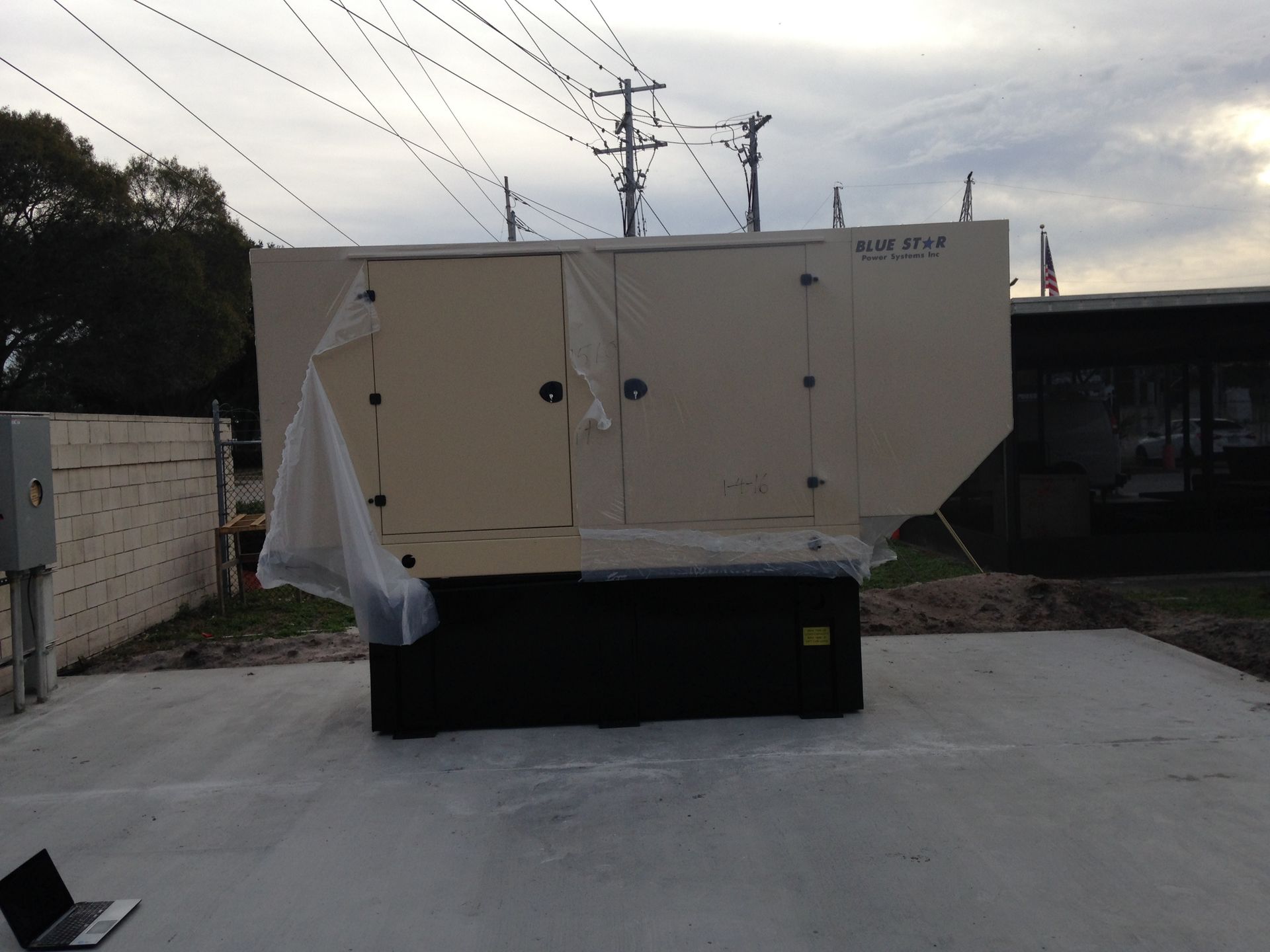 Large white industrial generator or machine on a driveway under power lines, with a black base and plastic cover.