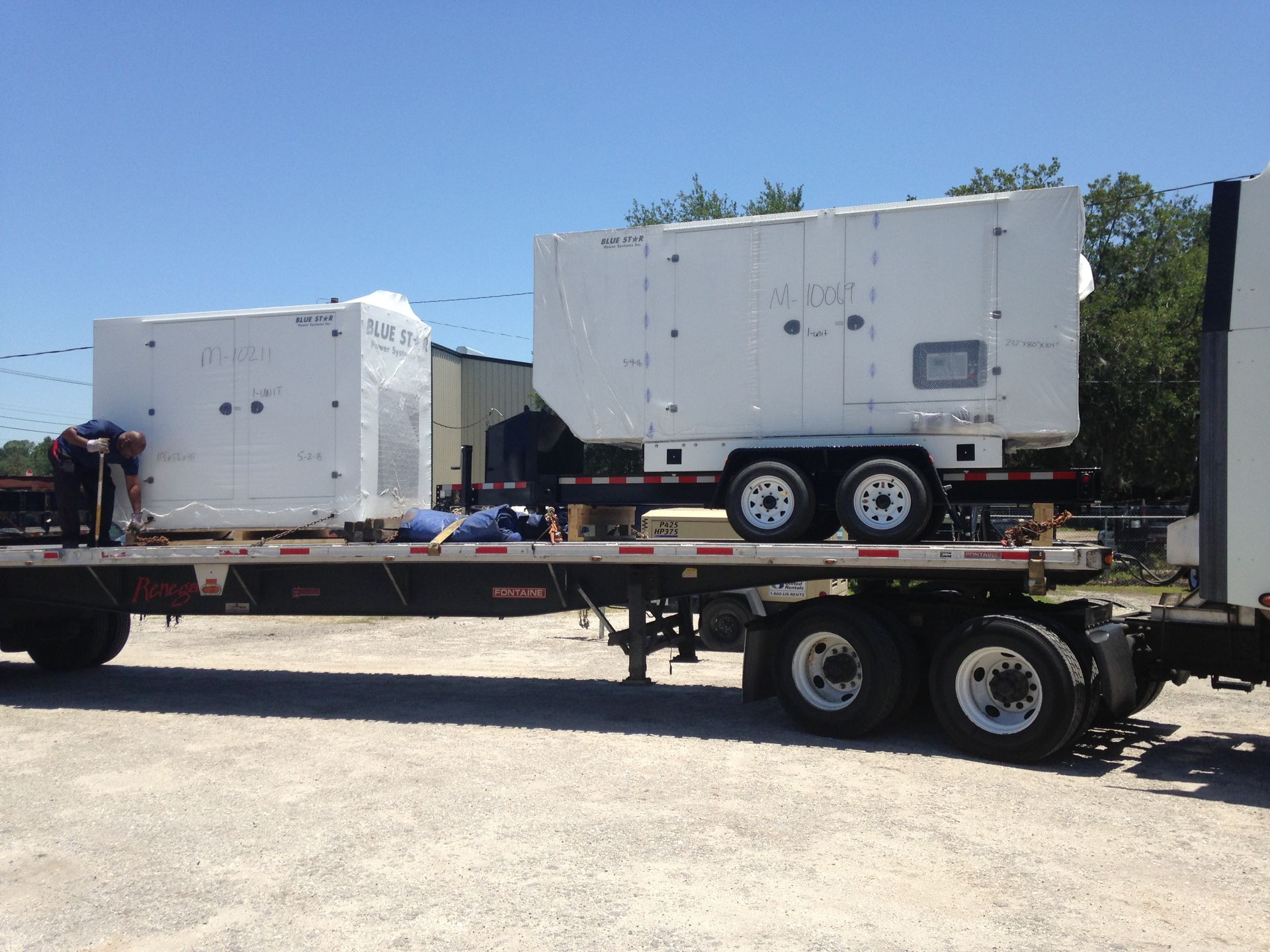 Two white industrial trailers loaded on a flatbed truck in a sunny yard