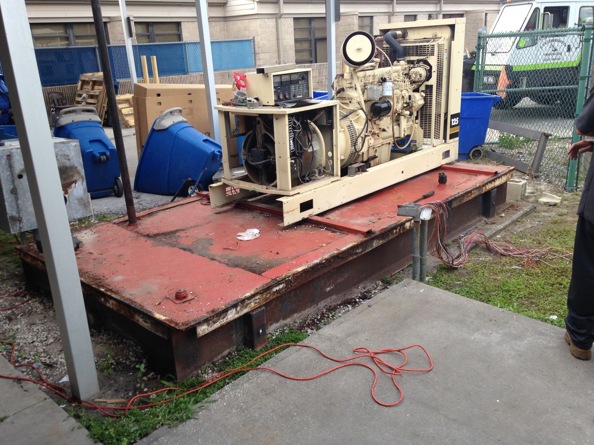 Industrial machinery on a red platform outdoors, with blue barrels and a chain-link fence nearby.