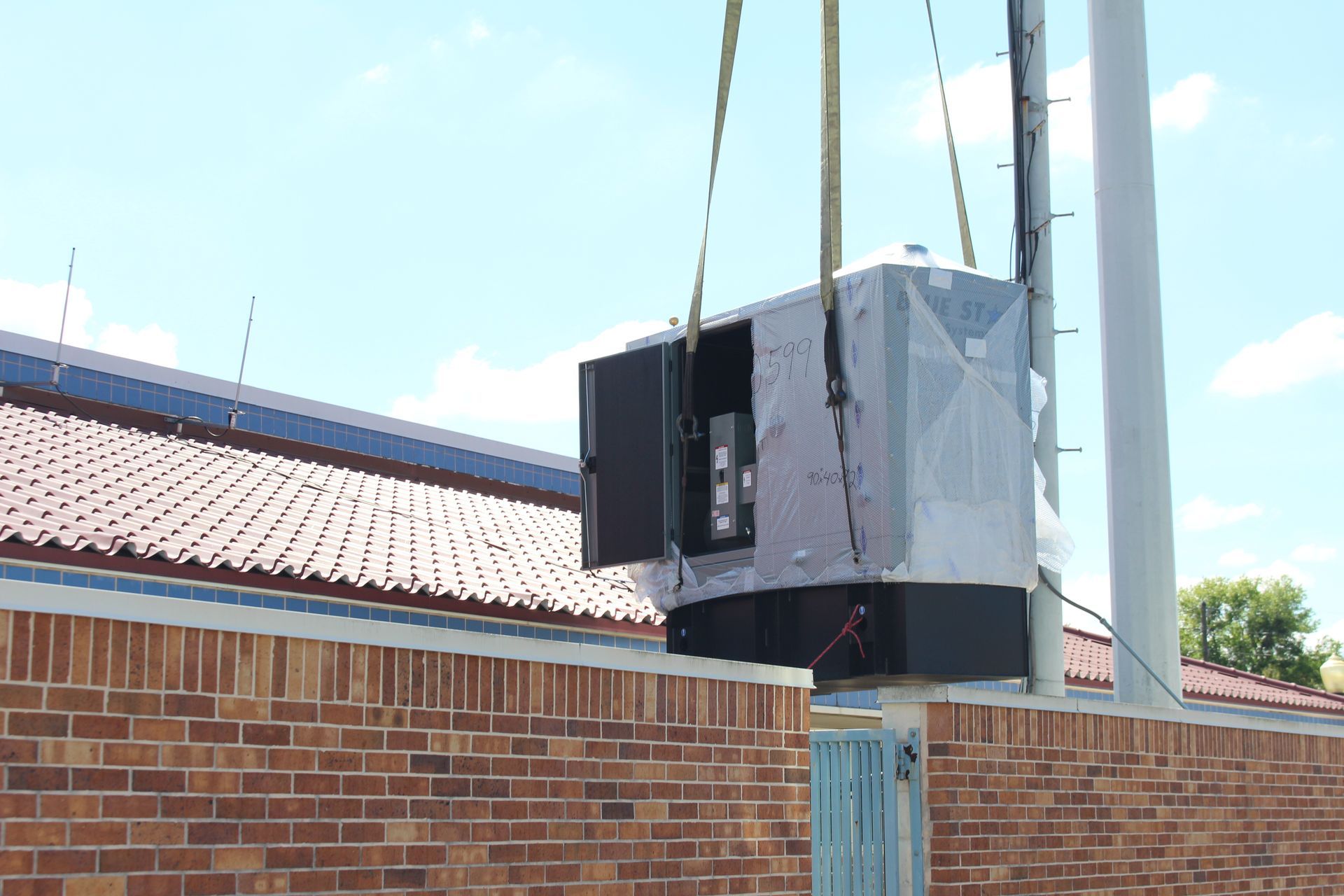 Large gray box lifted by crane beside a brick building under a blue sky