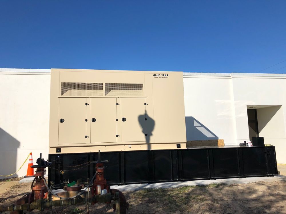 Beige industrial generator unit beside a white building, with black barriers and orange cones in front.