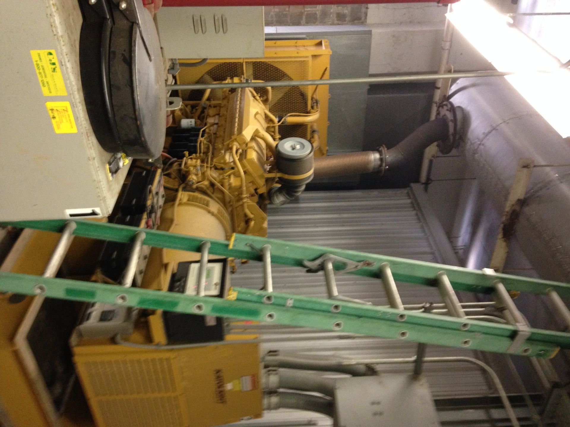 Industrial machinery and green ladder inside a mechanical room with metal ducts and pipes