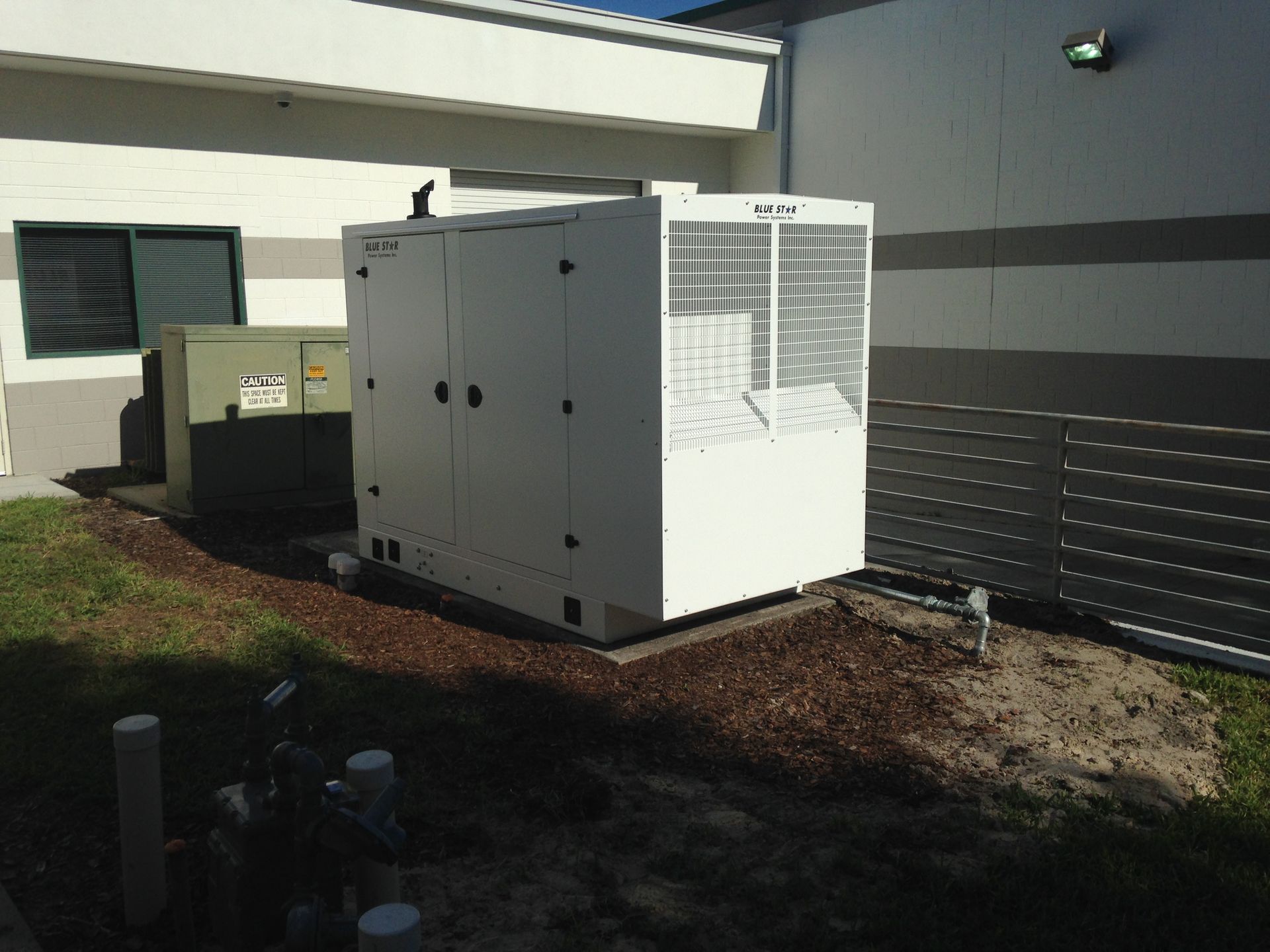 White generator unit beside a building on a gravel patch, with a fenced section and wall vents nearby